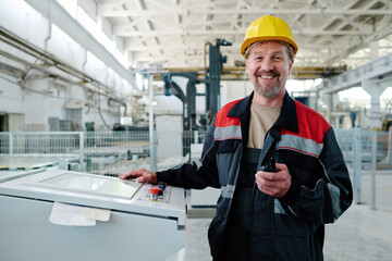 Mature engineer in helmet smiling at camera while controlling the work of production on machine