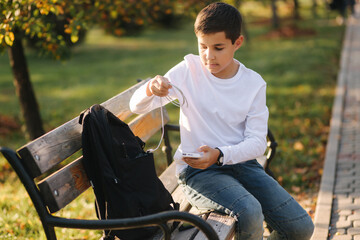 Handsome teenage boy take from his backpack powerbark for charging smarphone. Cute boy in the park in autumn time