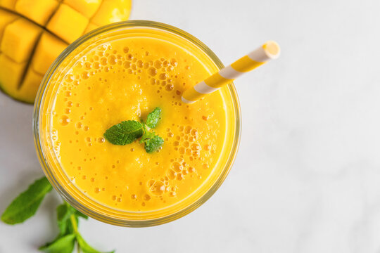 Fresh yellow summer mango smoothie in a glass with a straw and mint on white marble table. Top view
