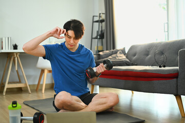 Active sportsman training arms with dumbbell and watching video online training on laptop at home
