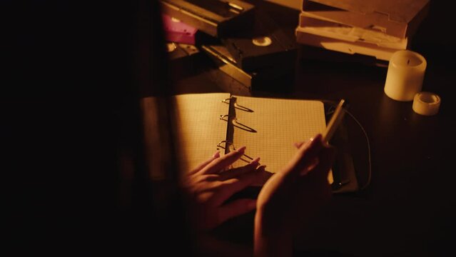 the girl flips through a notebook and writes in the dim light of a lamp