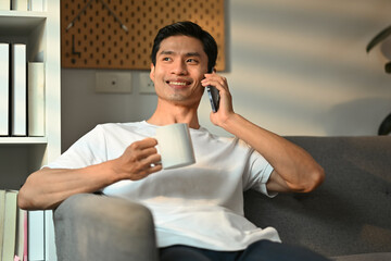 Positive millennial man having pleasant mobile phone conversation and drinking coffee in living room