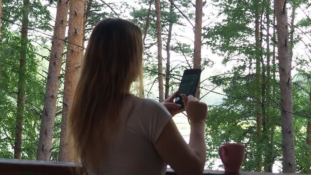 Happy woman relaxing and looking forest and lake at countryside home or homestay in the morning. drinks tea or coffee. taking photos and selfies. Vacation, blogger, SoloTravel, journey, trip