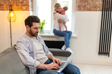 Fototapeta premium Bearded man working on computer, wife and toddler daughter on the background hugging.
