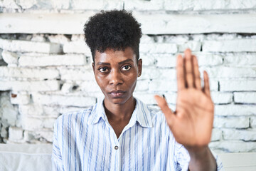 Obraz premium A miggle-aged african woman sitting on her sofa standing with outstretched hand showing stop sign, preventing you.