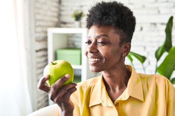 African woman in her living room, eating a green apple, embodying a healthy lifestyle.
