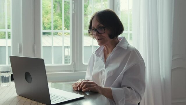 An elderly woman with dark hair in a white shirt sits at a round wooden table in front of a laptop and communicates via video link. Video call. Communication of people in the network.