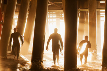 Surfers carrying boards under pier - obrazy, fototapety, plakaty
