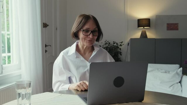 An elderly woman with dark hair in a white shirt sits at a round wooden table in front of a laptop and communicates via video link. Video call. Communication of people in the network.