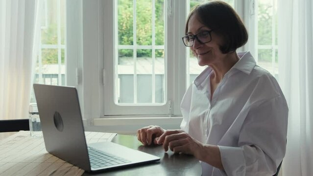 An elderly woman with dark hair in a white shirt sits at a round wooden table in front of a laptop and communicates via video link. Video call. Communication of people in the network.