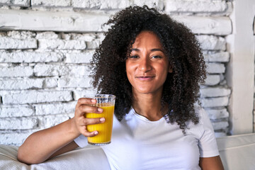 African-American woman enjoys freshly squeezed orange juice on sofa, morning light.