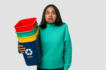 Young indian woman holding a recycling boxes cut out isolated shrugs shoulders and open eyes...
