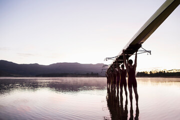 Rowing team entering lake at dawn with scull overhead 