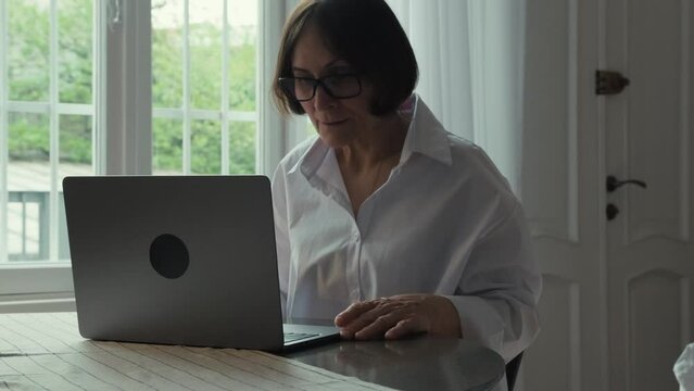An elderly woman with dark hair in a white shirt sits at a round wooden table in front of a laptop and communicates via video link. Video call. Communication of people in the network.
