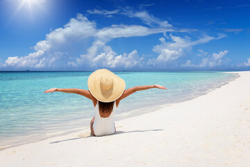 A happy woman in a white bathing suit sits on a tropical beach and enjoys her summer vacation time