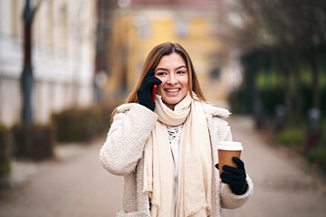 Young cheerful woman walking on city streets with hot coffe in her hand while talking on her mobile phone  - Outdoor smartphone usage concept in cold winter weather with a girl wearing jacket