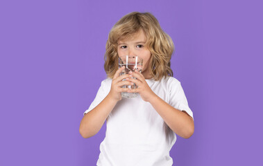 Little kid drinking a fresh glass of water, isolated on studio background. Thirsty child holding glass drinks still water. Portrait of kid boy drinking pure water from glass.