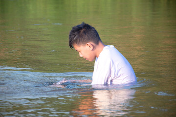 Asian boy in white t-shirt is spending his freetimes by diving, swimming, throwing rocks and catching fish in the river happily, hobby and happiness of children concept, in motion.