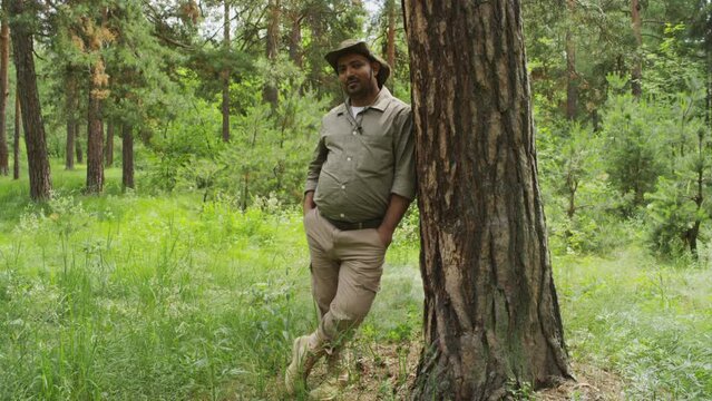 Full portrait shot of multiethnic man in khaki coloured forest warden uniform and sunhat standing in pine forest with hands in pockets, leaning on tree and looking at camera with confident smile