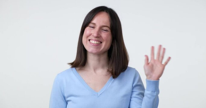 A cheerful Caucasian woman standing on a white background waves her hand in a welcoming gesture and flashes a warm smile, expressing her friendliness and good mood.