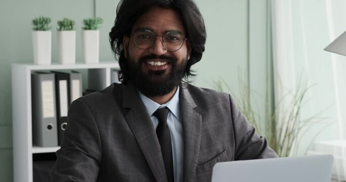 Slow-motion shot of an Indian CEO sitting in an office and looking directly into the camera with a big, toothy smile. Businessman radiating confidence as he presents a friendly and approachable image.
