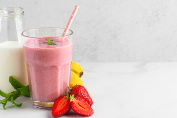 Strawberry and banana milkshake in a glass with a straw, fresh fruits and bottle of milk on white...
