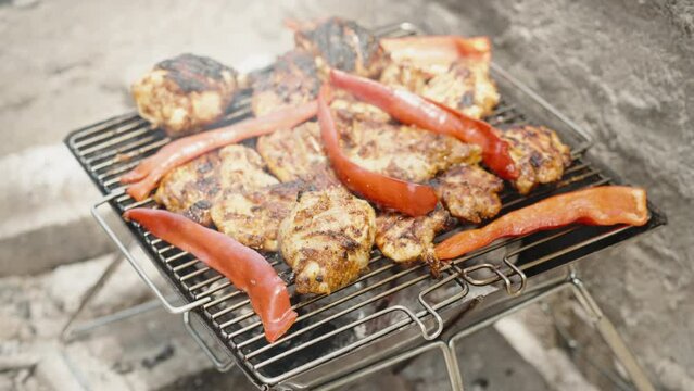 On the portable small grill, chicken is being grilled together with red sweet pepper, and smoke is coming out of them.