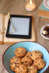Books, reading glasses, e-reader, plate of chocolate pralines, bowl of cookies, cups of tea and lit candles on the table. Selective focus.