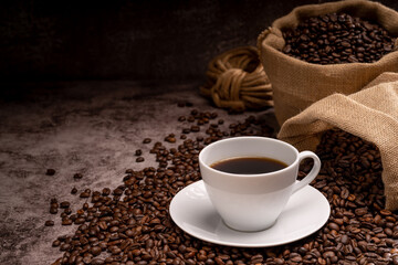 Hot coffee in a white coffee cup and many coffee beans placed around on a wooden table in a warm, light atmosphere, on dark background.