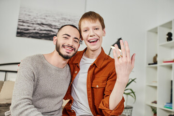 carefree gay man showing wedding ring near smiling bearded boyfriend at home.