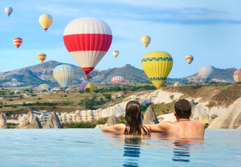 Romantic couple watches colorful balloons flying over mountains on sunny morning in luxury infinity pool.