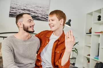 cheerful redhead gay man showing wedding ring near bearded boyfriend at home.