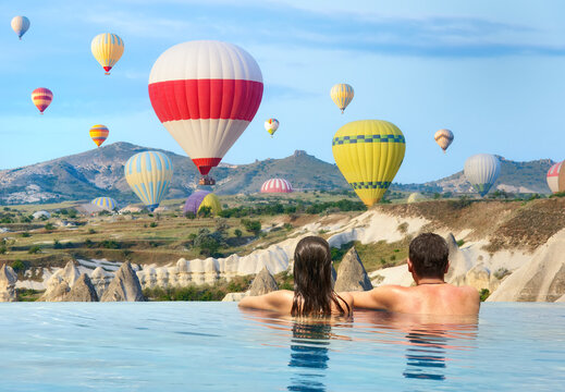 Romantic couple watches colorful balloons flying over mountains on sunny morning in luxury infinity pool. - Powered by Adobe
