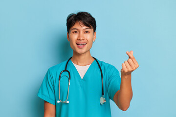 Kind young doctor stands on blue background, folding fingers of his left hand in heart gesture, smiling at camera, professional people concept, copy space