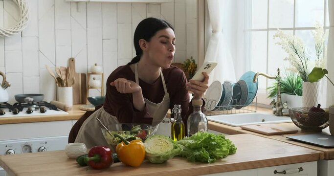 Caucasian woman wearing a kitchen apron stands near a table in a cozy kitchen. With a phone in hand, she effortlessly multitasks, browsing through her device while relishing a fresh and vibrant salad.