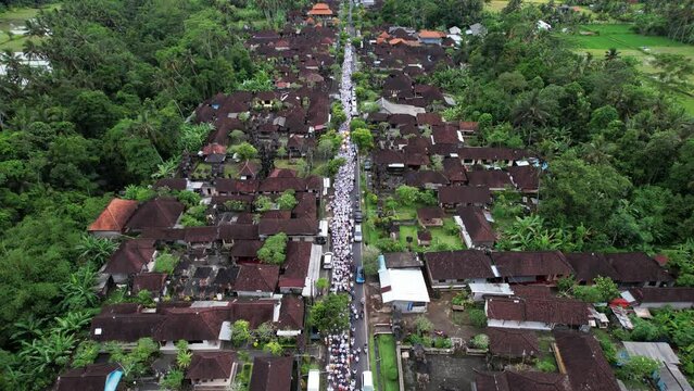 Huge religious procession walk at village street, aerial lengthwise view, camera fly forward. Melis day celebration at Bali Island, people walk together to beach, carry offerings, wearing white