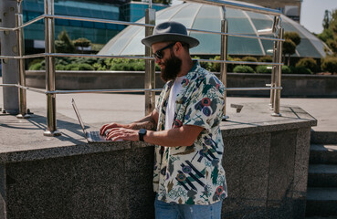 Photo of a freelance web designer young guy working outdoors on a laptop computer connected to a public Wi-Fi. Carefree caucasian hipster spends his free time in the park.