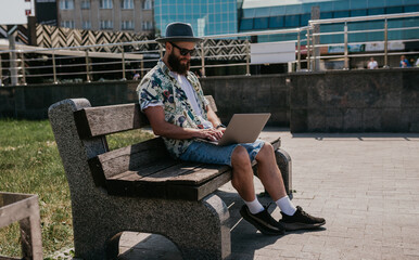 Photo of a freelance web designer young guy working outdoors on a laptop computer connected to a public Wi-Fi. Carefree caucasian hipster spends his free time in the park.