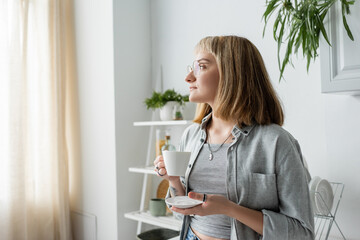 young woman with short hair and bangs, eyeglasses and tattoo on hand holding cup of morning coffee and saucer while standing in grey casual clothes next to dishes rack and plant in modern kitchen - obrazy, fototapety, plakaty