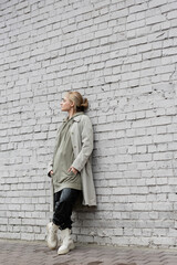 young woman with makeup, blonde hair, bangs, in stylish outfit with long hoodie, coat, black leather pants and beige boots standing with closed eyes near grey brick wall of modern building