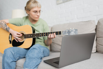 blurred young woman in glasses with bangs and tattoo playing acoustic guitar and looking video tutorial on laptop while sitting on comfortable couch in modern living room, virtual learning