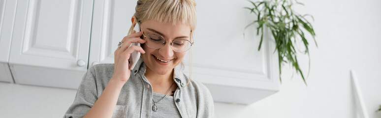 happy young woman with bangs and eyeglasses smiling and talking on smartphone while standing with closed eyes near blurred green plants and white kitchen cabinets in modern apartment, banner