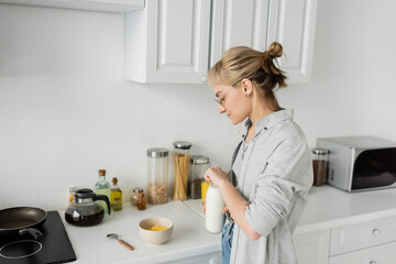 young woman in eyeglasses and short hair with bangs holding bottle with fresh milk near bowl with cornflakes while making breakfast and standing in casual clothes next to kitchen appliances at home