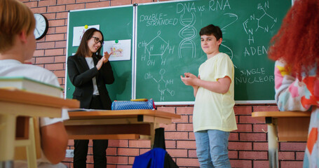 Friendly female teacher of elementary school listening attentively schoolboy answer about DNA structure near blackboard. Biology lesson for pupils at class.