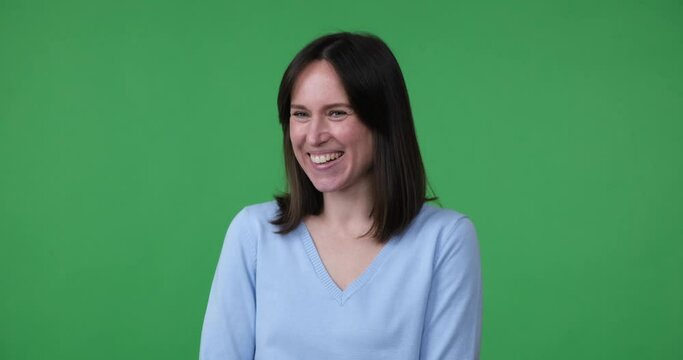 A shy Caucasian woman stands on a green screen background, smiling and covering her face with her hand in a bashful manner. She looks very embarrassed.