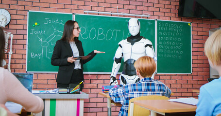 Caucasian female teacher with tablet in hands presenting humanoid robot to attentive pupils at...