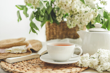Cup of tea and teapot on windowsill and basket with white lilacs, good morning concept