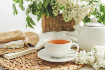 Cup of tea and teapot on windowsill and basket with white lilacs, good morning concept