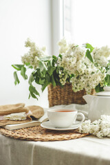 Cup of tea and teapot on windowsill and basket with white lilacs, good morning concept