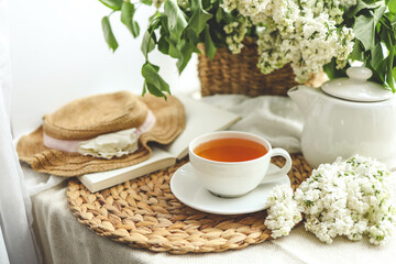 Cup of tea and teapot on windowsill and basket with white lilacs, good morning concept